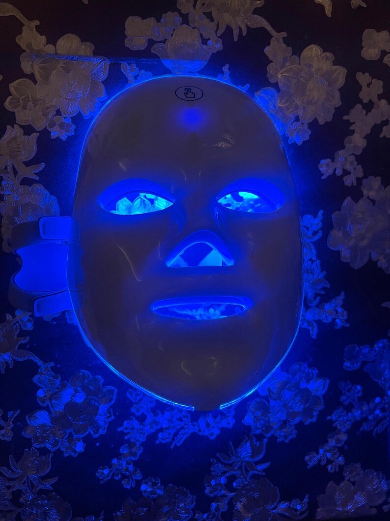 May include: A white LED light therapy mask with blue illumination around the eyes, nose, and mouth. The mask is designed to cover the entire face. The background is a dark blue floral pattern.