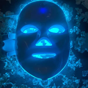 May include: A dark blue LED face mask with glowing blue lights around the eyes, nose, and mouth. The mask has adjustable straps and is designed to fit the contours of the face. The background features a dark surface with a floral pattern.
