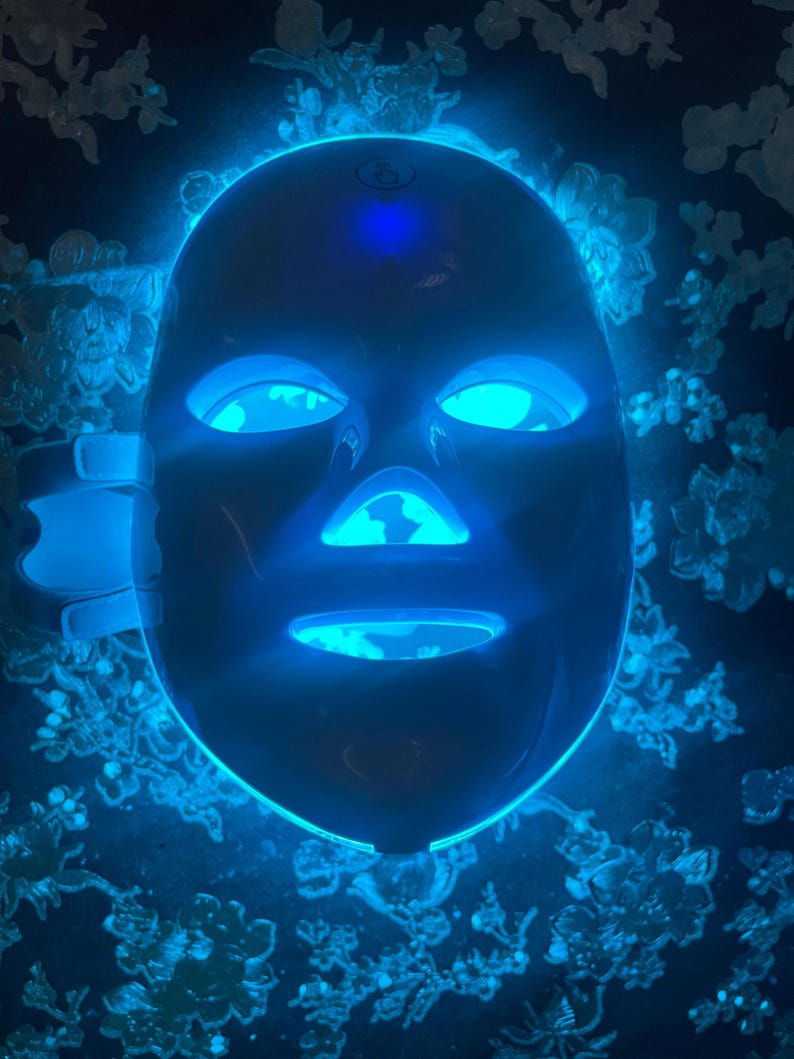 May include: A dark blue LED face mask with glowing blue lights around the eyes, nose, and mouth. The mask has adjustable straps and is designed to fit the contours of the face. The background features a dark surface with a floral pattern.