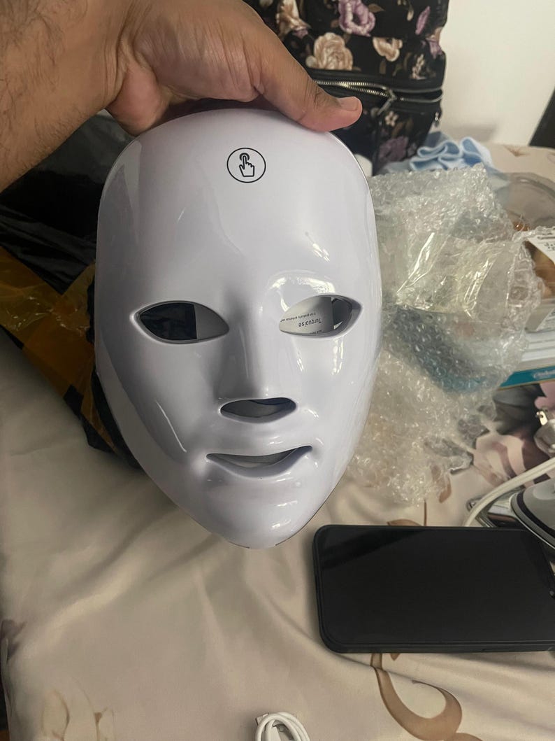 May include: A white LED face mask with eye, nose, and mouth openings. The mask has a touch-sensitive power button and is held by a hand. A black smartphone and other items are in the background.