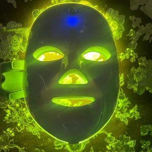 May include: A white LED light therapy mask with green and yellow lights illuminating the eye, nose, and mouth openings. The mask has a blue light on top and a green strap on the side. The background is a dark color with a floral pattern.