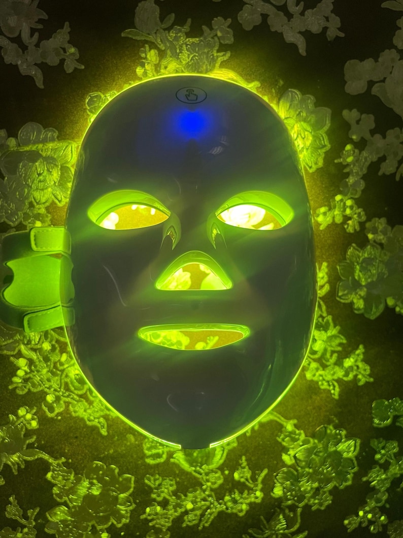 May include: A white LED light therapy mask with green and yellow lights illuminating the eye, nose, and mouth openings. The mask has a blue light on top and a green strap on the side. The background is a dark color with a floral pattern.