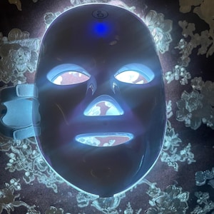 May include: A white LED light therapy mask with blue and white lights. The mask has openings for the eyes, nose, and mouth, and adjustable straps. The mask is designed for skincare and beauty treatments.