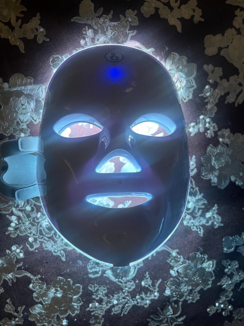 May include: A white LED light therapy mask with blue and white lights. The mask has openings for the eyes, nose, and mouth, and adjustable straps. The mask is designed for skincare and beauty treatments.