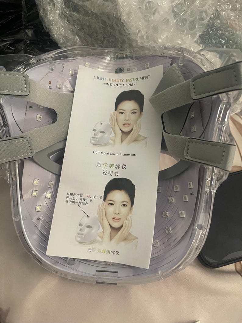 May include: A clear, light therapy face mask with gray adjustable straps. The mask has numerous small LED lights and includes a printed instruction sheet with text and illustrations. The text on the sheet reads "LIGHT BEAUTY INSTRUMENT".