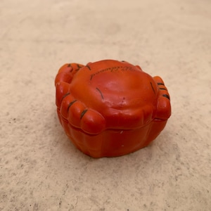 May include: Orange ceramic crab trinket box with black detailing. The box is closed and the top of the box has the word "Switzerland" written on it.