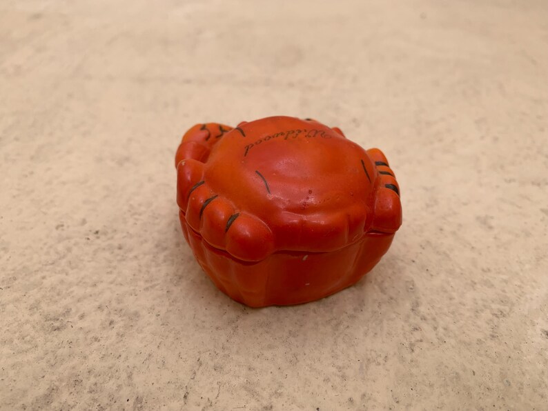 May include: Orange ceramic crab trinket box with black detailing. The box is closed and the top of the box has the word "Switzerland" written on it.