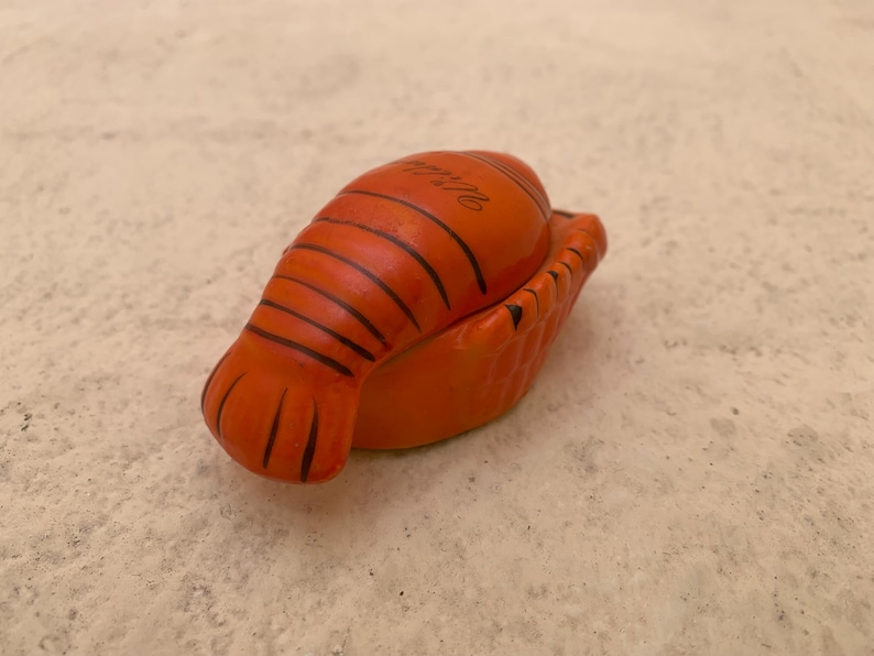 May include: Orange ceramic lobster shaped trinket box with black detailing. The box is closed and the top of the box has the text "Italia" written on it.