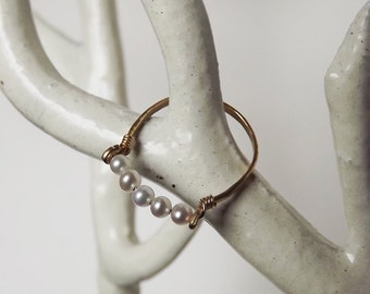 May include: A gold ring with a row of small white pearls. The ring is displayed on a white ceramic stand.