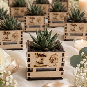 May include: A square wooden planter box containing a green succulent plant. The front panel is laser-engraved with the text Stella & Felix and the date 21st Jan 2024, accompanied by a line-art illustration of a couple in wedding attire.