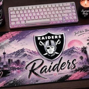 以下が含まれることがあります： ラスベガス・レイダースのロゴ、「Raiders」と「Just Win Baby」の文字、山々、ヤシの木、大きな月が描かれたピンク調の都市風景が特徴の長方形のデスクマット。デザインには筆致のアクセントと小さなハートのアイコンが含まれています。