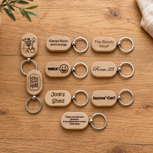 May include: A collection of light-coloured wooden oval keychains with metal rings. The keychains feature various laser-engraved designs and text including "Games Room and Lounge," "The Bloody Keys!," "SMILE!," "Room 23," "Jodi's Shed," "James' Car!," "HOME SWEET HOME," and a gardening icon set.