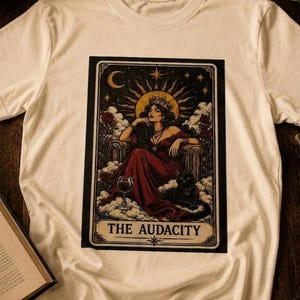 May include: A cream-colored short-sleeve t-shirt featuring a rectangular graphic print of a person in a red dress wearing a crown, seated on a throne with a black cat and a wine glass. The text THE AUDACITY is printed at the bottom of the graphic.