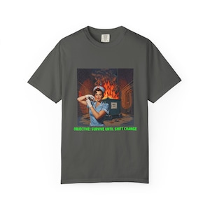 Op de afbeelding: Donkergrijs t-shirt met een retro-illustratie van een verpleegster in een blauw uniform, tegen een achtergrond van een brandende vuilnisbak. De groene tekst luidt "OBJECTIVE: SURVIVE UNTIL SHIFT CHANGE".