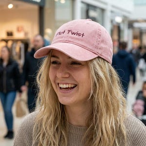Puede incluir: Una gorra de béisbol color rosa empolvado con el texto Plot Twist bordado en hilo marrón oscuro en el panel frontal. La gorra tiene una visera curva y una corona estructurada con un botón en la parte superior, hecha de algodón suave lavado.