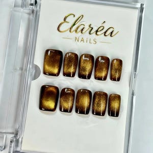 Peut inclure: Un ensemble de dix faux ongles carrés dans un étui en plastique transparent. Les ongles présentent un dégradé, passant du marron foncé à la base au jaune doré en haut. La marque "Elaréa Nails" est affichée en lettres dorées.