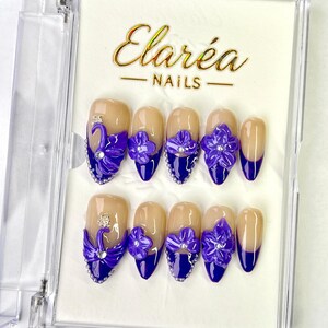 Pode incluir: Um conjunto de unhas postiças numa caixa transparente. As unhas apresentam uma base nude com uma ponta azul escura. Cada unha é adornada com uma flor roxa e strass prateados. A marca "Elaréa Nails" é exibida em letras douradas.