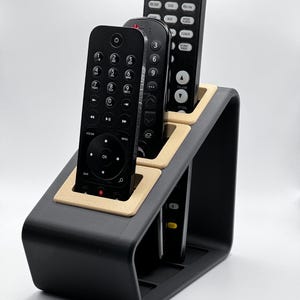 May include: A black remote control holder with a slanted design and light tan interior accents. The holder features three vertical slots, each containing a black remote control with various buttons, including numeric keys and directional pads.