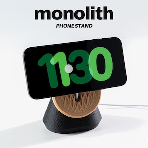 Può includere: Uno smartphone nero che mostra l'ora 11:30 in verde è appoggiato su un supporto per telefono nero e color legno. Il supporto ha un design geometrico ed è collegato a un cavo di ricarica bianco. Le parole "monolith" e "PHONE STAND" sono stampate sopra il telefono.