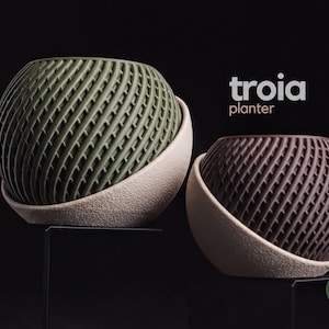 May include: Two Troia planters with a spherical, textured grid pattern. One planter is olive green and the other is dark brown, both set within a light beige, rough-textured, semi-circular base. The text "troia planter" appears in white.