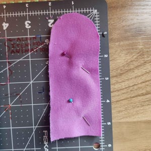 May include: A piece of purple fabric with a rounded top edge, secured to a black Fiskars cutting mat with two sewing pins. The fabric measures approximately 5.5 inches in length and 2 inches in width, as indicated by the grid markings.