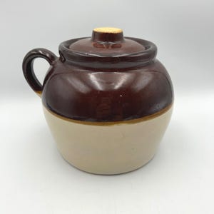 May include: A round ceramic crock with a dark brown glazed upper section and a light beige lower section. The vessel features a single side handle, a matching dark brown lid with a small knob, and a smooth, glossy finish on the top half.