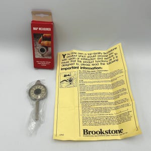 May include: A Brookstone map measurer in a red cardboard box, a silver-toned metal map meter inside a clear plastic bag, and a yellow instruction sheet with black text titled Important Information: No. 2962 Map Meter (Opisometer).