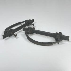 May include: Two dark gray metal drawer pulls with a semi-circular handle design. Each pull features a rectangular mounting plate with two protruding metal pins and a curved, hinged handle that swings downward from the central horizontal bar.