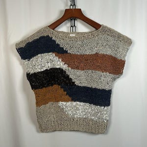 May include: A sleeveless, open-knit top with a crew neck. The top features horizontal stripes in shades of beige, navy, brown, black, and silver. The garment is hanging on a wooden hanger against a white background.