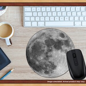 May include: A round mousepad featuring a detailed grayscale image of the moon. A black wireless mouse sits to the right of the mousepad. A white keyboard, a cup of coffee, a wallet, and a pencil are also on the wooden desk.