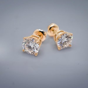 May include: A pair of gold stud earrings featuring round, clear, faceted stones held in place by four-prong settings. The earrings have a polished gold finish and are designed with a classic post back mechanism for secure attachment to the earlobe.