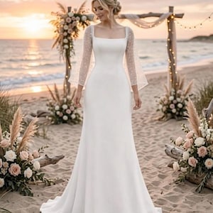 May include: A white wedding dress featuring a square neckline and sheer sleeves adorned with small dots. The dress has a fitted bodice and a flowing skirt. The image is set on a beach, with floral decorations and a wooden arch in the background.
