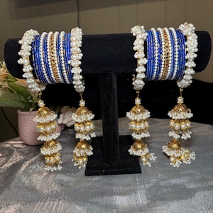 May include: Two sets of blue and white bangles with gold accents and dangling gold and pearl beaded ornaments. The bangles are displayed on a black stand, showcasing the intricate details of the jewelry.