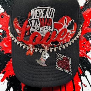 May include: A black trucker hat featuring a red and white embroidered patch with the text "WE'RE ALL MAD HERE" and a top hat icon. The hat includes a red script "Love" applique, a rhinestone diamond shape, and a pearl chain with a top hat charm.