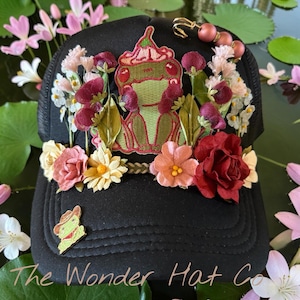 May include: A black baseball cap adorned with a central embroidered green frog patch, surrounded by various artificial fabric flowers in shades of red, pink, and white. A small frog pin and a beaded safety pin are attached to the cap.