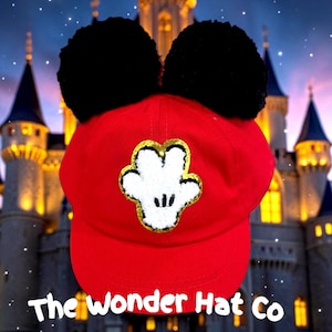 May include: A bright red baseball cap featuring two large black pom-poms on top and a central white glove patch with a gold glitter border. The text The Wonder Hat Co is printed in white at the bottom against a castle background.