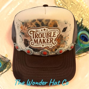 May include: A brown and cream trucker hat featuring a central graphic with the text TROUBLE MAKER, surrounded by a floral and leopard print pattern. The hat is accented with a silver-toned metal band containing small, oval-shaped red stones.