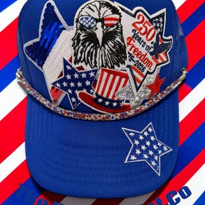 May include: A royal blue trucker hat featuring a central patch of an eagle wearing flag-patterned sunglasses, a top hat, and star-shaped patches. The hat is accented with a red, white, and blue rhinestone trim and includes the text 250 Years of Freedom 1776-2026.