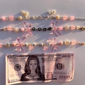 May include: Three pink beaded bracelets with the name "KALI" spelled out in black and white letter beads. The bracelets feature pink bows and various beads. A one hundred dollar bill is in the foreground.