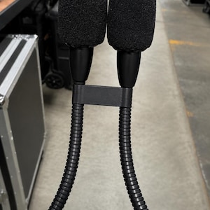 Puede incluir: Dos micrófonos negros con cubiertas de espuma, conectados por un soporte negro y tallos flexibles y acanalados. Estos micrófonos son probablemente para grabación de audio profesional o actuaciones en vivo. El fondo muestra estanterías y estuches.
