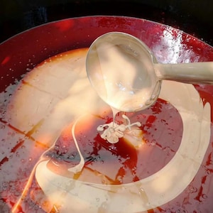 May include: A metal ladle pours a thick, creamy beige liquid into a large basin filled with a deep, translucent red liquid. The beige substance creates swirling patterns and textured drips as it merges with the dark red base, creating a contrast.