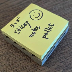 Op de afbeelding: Een gele 7,6 x 7,6 cm plaknotitieblok met een smiley en de woorden "Sticky notes pallet" in zwarte inkt. Het blok zit in een grijze plastic houder.