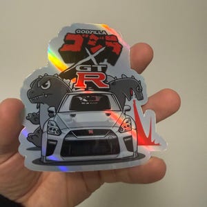 May include: A holographic sticker featuring a white Nissan GT-R sports car centered between two cartoon Godzilla figures. The text GODZILLA is printed in black above the red GT-R logo. The design includes a red beam effect on the right side.