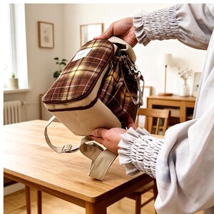 May include: A small backpack featuring a brown and yellow plaid pattern on the upper section and a solid beige base. The bag includes a white rectangular label with text on the front, a zipper closure, and adjustable beige shoulder straps.