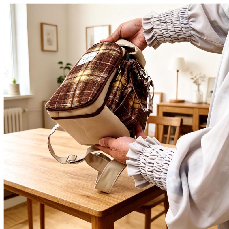 May include: A small backpack featuring a brown and yellow plaid pattern on the upper section and a solid beige base. The bag includes a white rectangular label with text on the front, a zipper closure, and adjustable beige shoulder straps.