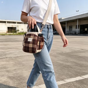 May include: A crossbody bag featuring a brown and tan plaid pattern on the front pocket, a cream-colored body, and a wide adjustable shoulder strap. The bag includes a side pocket and a small rectangular white label with black text on the front.
