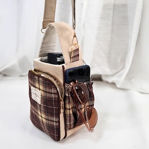 May include: A beige and brown plaid crossbody bag featuring a front zippered pocket, a side pocket holding a smartphone and sunglasses, and a top handle. The bag includes a rectangular fabric label with text and an adjustable shoulder strap.