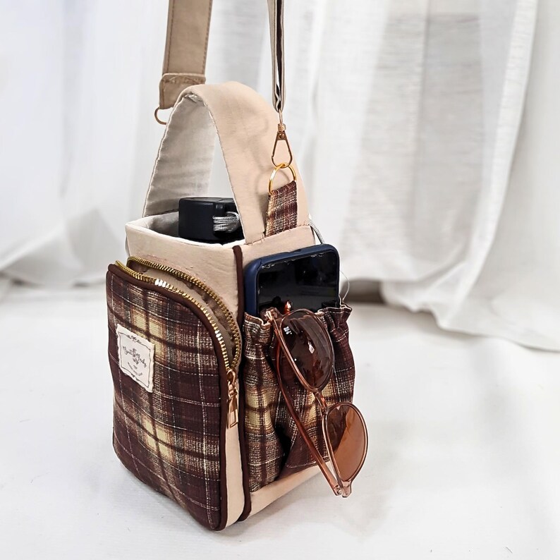 May include: A beige and brown plaid crossbody bag featuring a front zippered pocket, a side pocket holding a smartphone and sunglasses, and a top handle. The bag includes a rectangular fabric label with text and an adjustable shoulder strap.