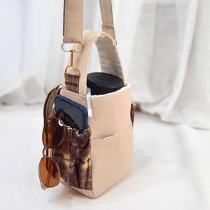 May include: A beige crossbody bag with a plaid fabric side pocket containing a dark smartphone and brown-tinted sunglasses. The bag features a top handle, an adjustable shoulder strap with gold-tone hardware, and a black cylindrical container inside.