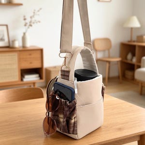 May include: A beige crossbody bag with a top handle and an adjustable strap. The bag features an exterior pocket with a brown and beige plaid pattern. A dark blue smartphone and brown-tinted sunglasses are tucked into the plaid pocket.